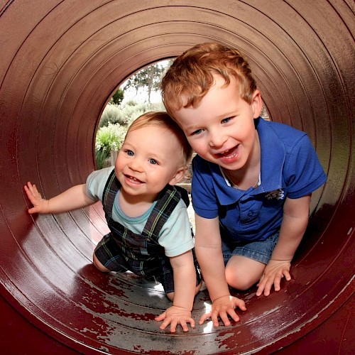 Crawling baby and young boy venturing out of a play tunnel, smiling